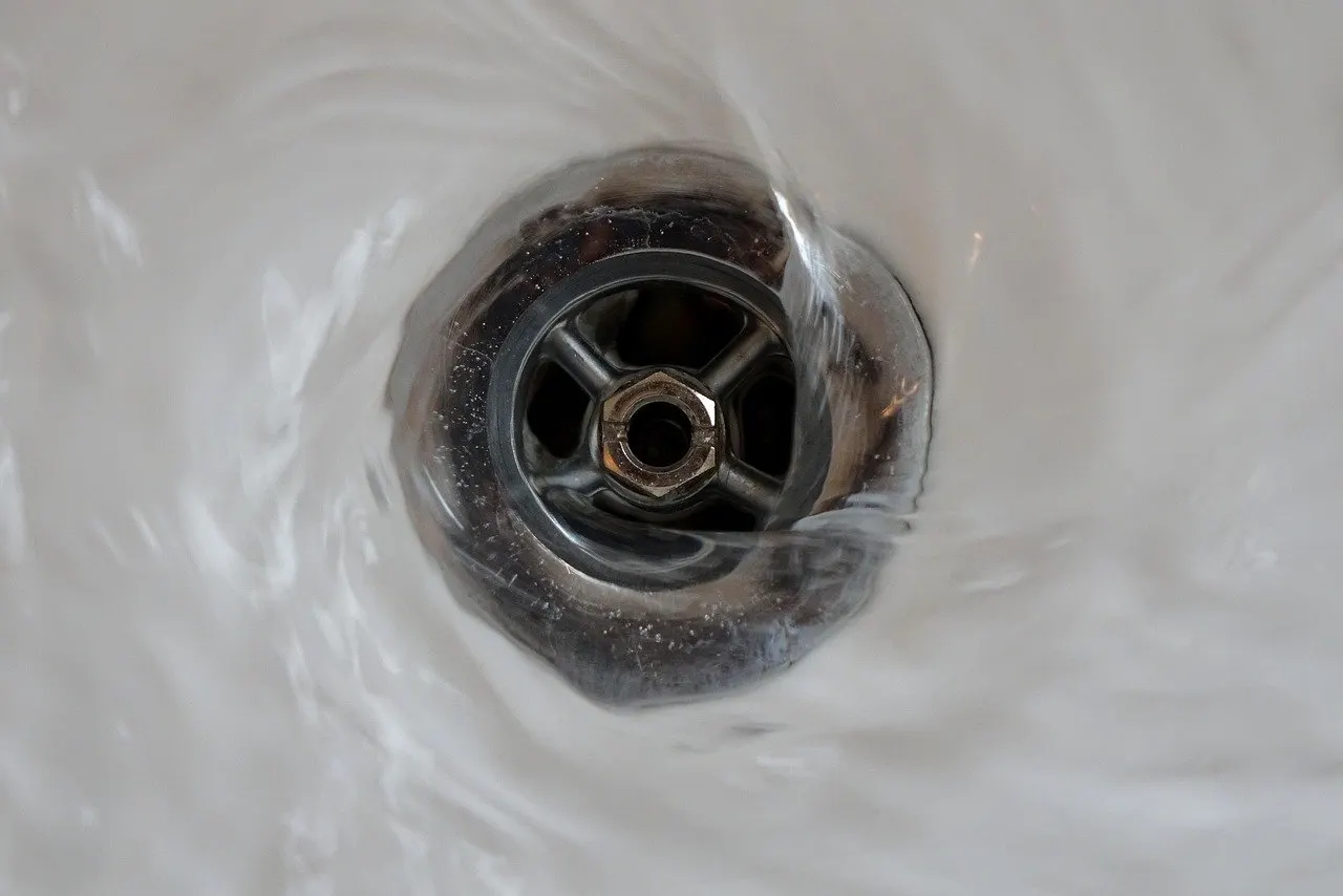 Wasser strudelt in den verchromten Ablauf eines weißen Beckens, unter dem Wasserfilm sind Ablaufsieb und Mittelschraube erkennbar