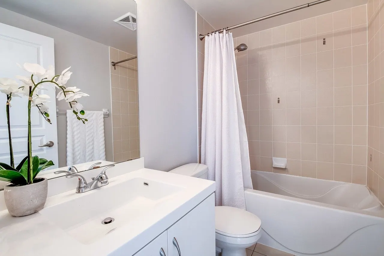 Kleines Bad mit Badewanne, Duschvorhang, WC und Waschtisch, im Wandspiegel ist ein Abluftgitter an der Decke zu sehen