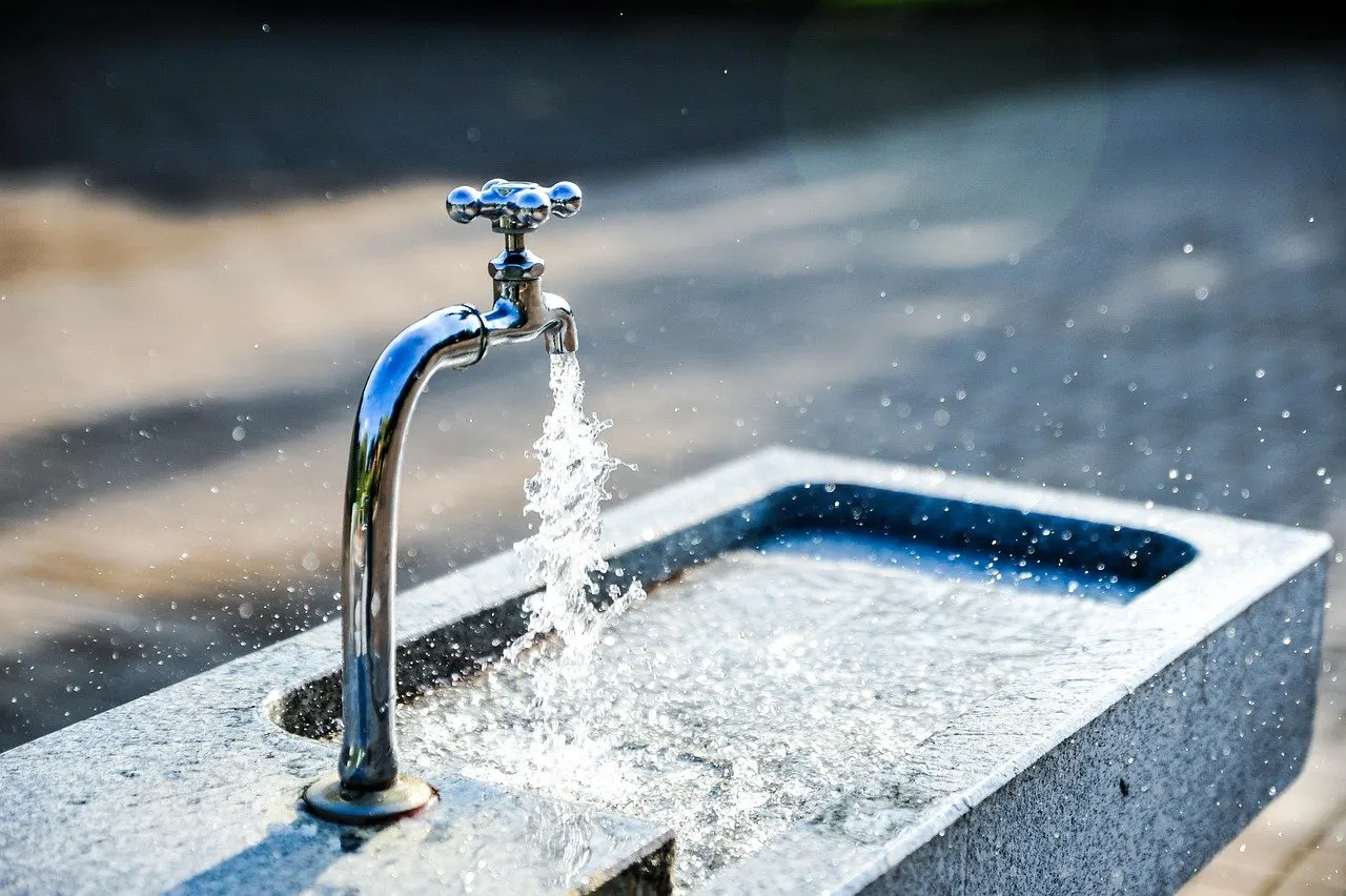 Verchromter Wasserhahn an einem Steinbecken im Freien, aus dem ein kräftiger Wasserstrahl spritzt, Hintergrund unscharf