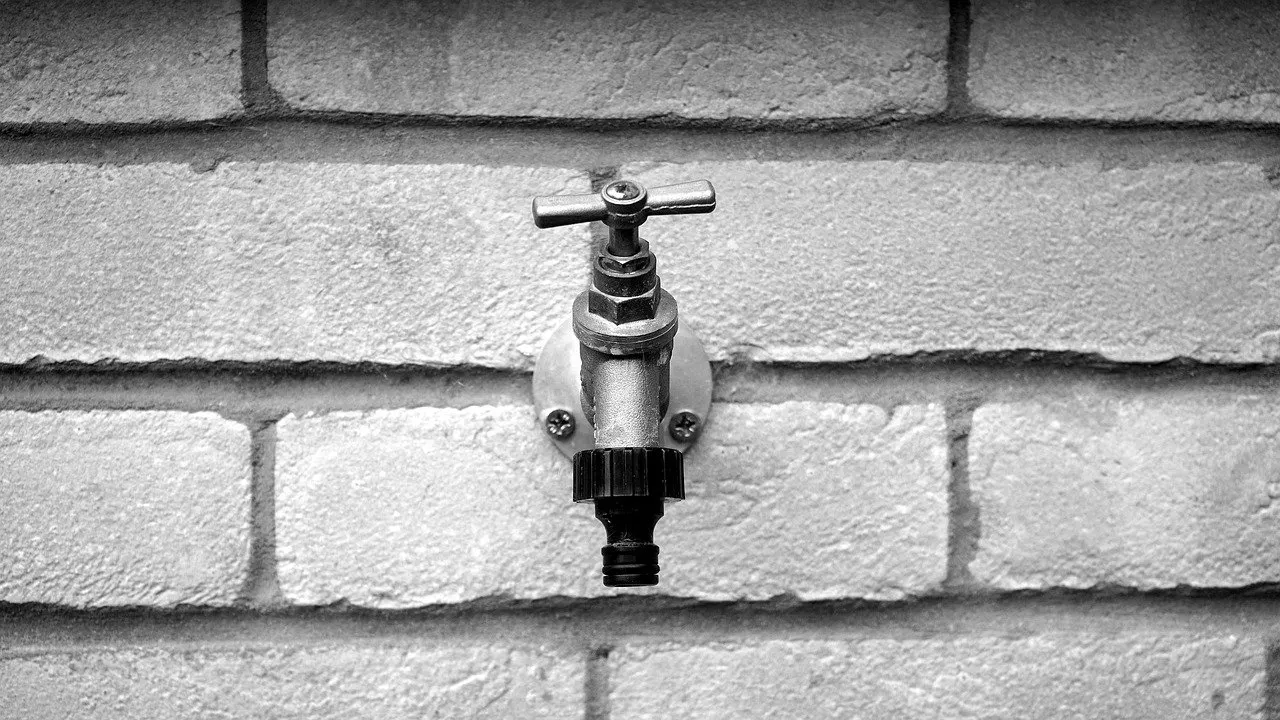Außenwasserhahn mit Knebelgriff und Schlauchanschluss an einer hellen Ziegelwand, Schwarzweißaufnahme