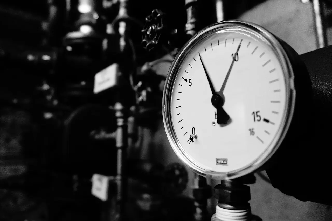 Schwarz-weiß-Aufnahme eines runden Manometers mit Skala bis 16 bar an einem Rohr, dahinter unscharf Ventile im Technikraum