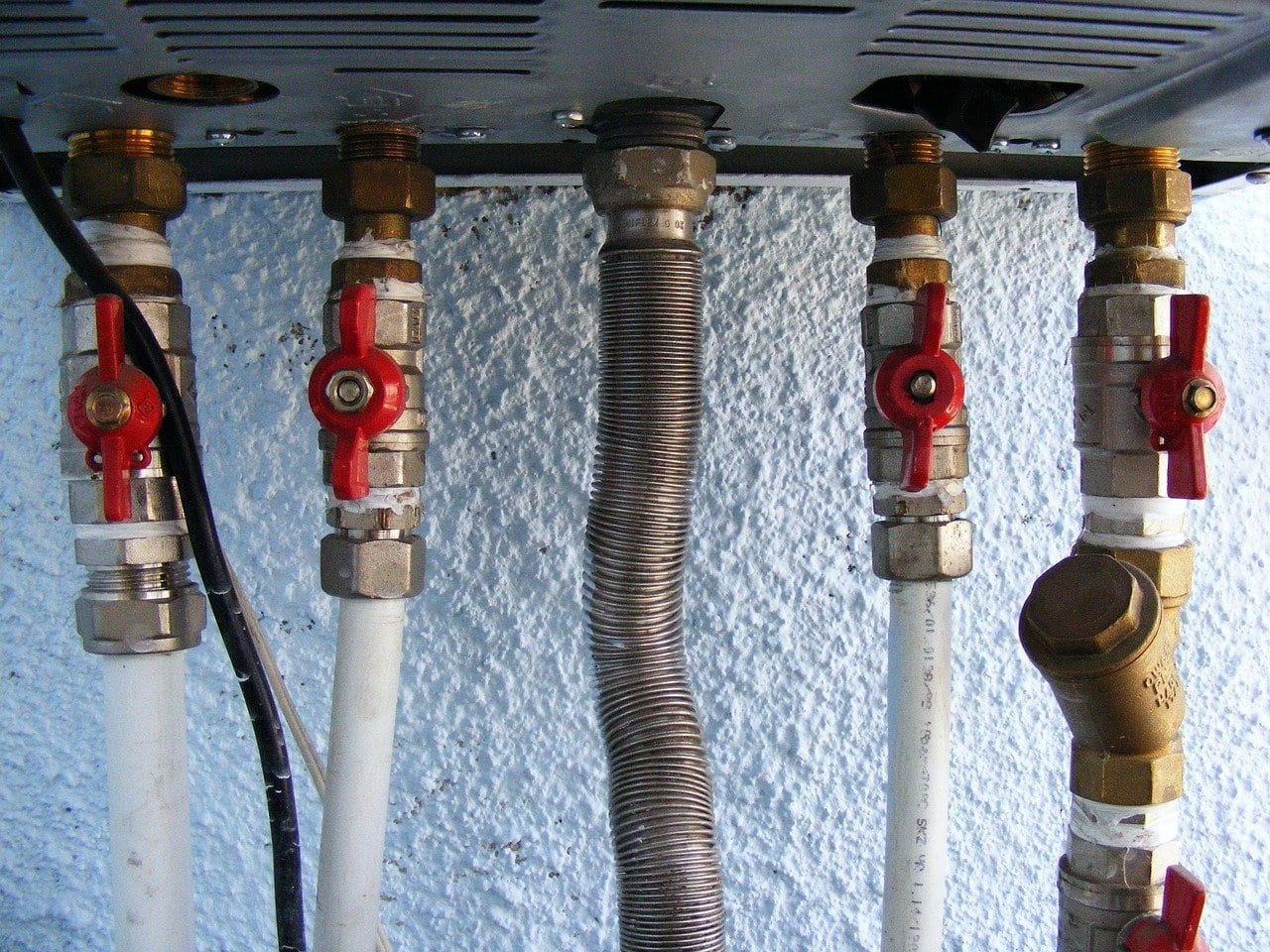 Anschlussleitungen unter einem Wandheizgerät mit roten Kugelhähnen, einem gewellten Edelstahlrohr und einem Schmutzfänger aus Messing