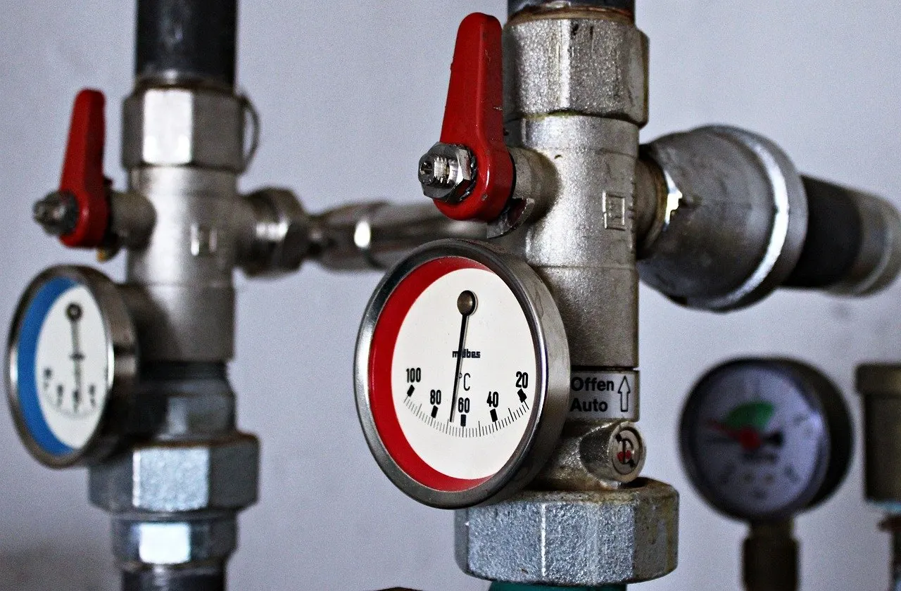 Vor- und Rücklaufleitung einer Heizungsanlage mit Zeigerthermometern, roten Kugelhähnen und einem Manometer im Hintergrund
