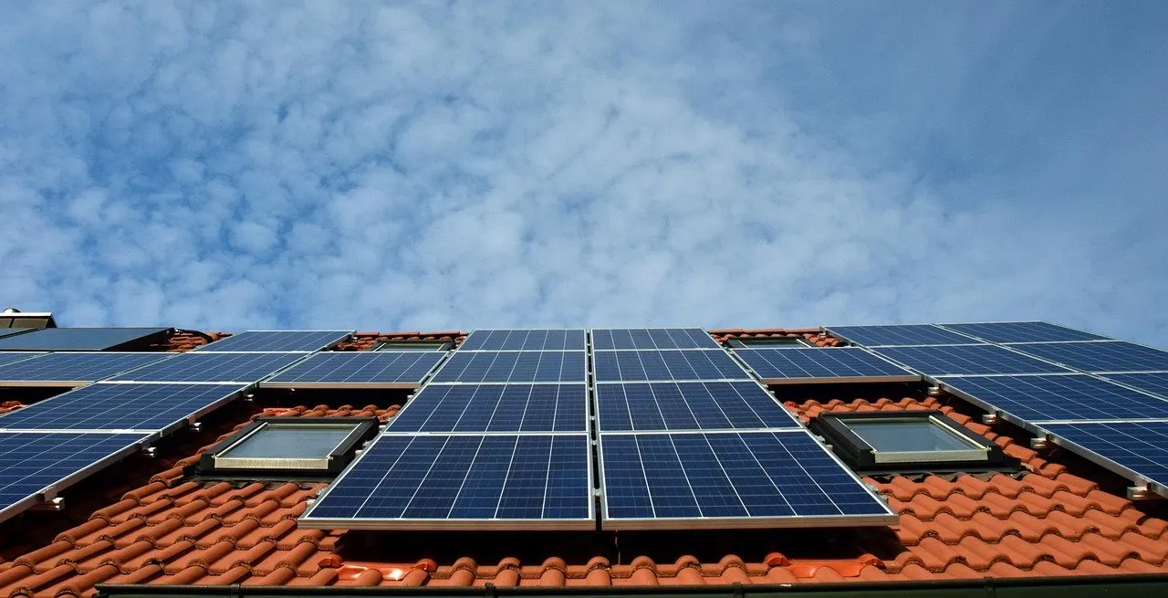 Solarmodule auf einem Ziegeldach mit roten Dachpfannen und zwei Dachfenstern, von unten fotografiert vor blauem, leicht bewölktem Himmel